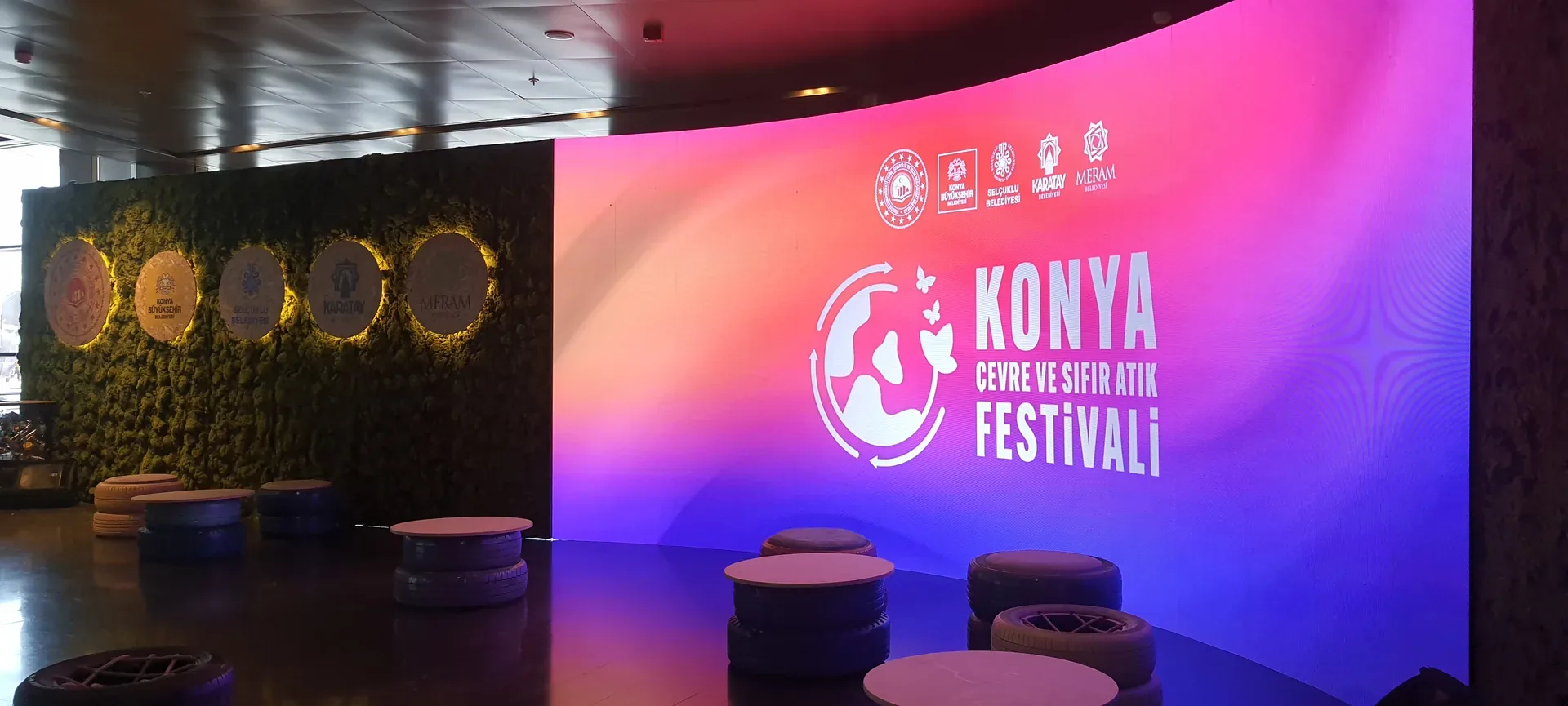 Festival fuayesinde kavisli LED ekran, yeşil duvar ve lastiklerden yapılan oturma grupları.